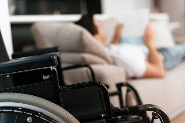 A fogyatékkal élő személy fekszik a kanapén, és olvas valamit a tabletta. A közelben van a kerekesszék. — Stock Fotó