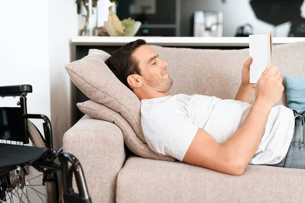 Osoba niepełnosprawna leży na kanapie i czyta coś na swoim tabletem. W pobliżu hotelu jest jego wózek inwalidzki. — Zdjęcie stockowe