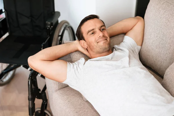Инвалид лежит на диване и отдыхает, положив руки за голову. Рядом его инвалидное кресло. . — стоковое фото