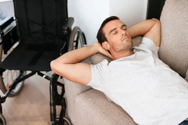 장애인 소파와 나머지에 그의 손을 그의 머리의 뒤에 누워 있다. 근처 그의 휠체어에는. — 스톡 사진