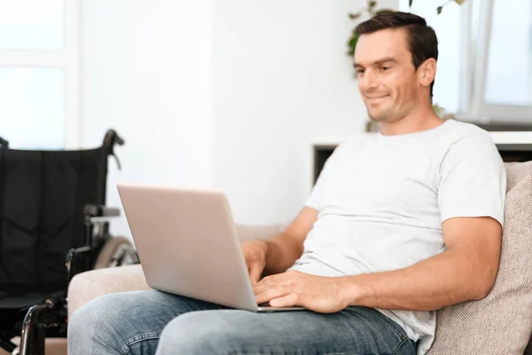 The disabled person is sitting on the couch. He works for his laptop. next is his wheelchair. — Stock Photo, Image