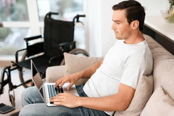 Den funktionshindrade personen sitter på soffan. Han arbetar för sin laptop. Nästa är hans rullstol. — Stockfoto