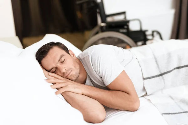 The disabled person sleeps in his white bed. He's sleeping. Behind him is his wheelchair. — Stock Photo, Image
