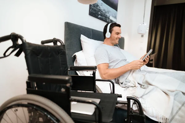 El inválido yace en la cama y se ve algo en la tableta. Él sonríe. porque es feliz . — Foto de Stock