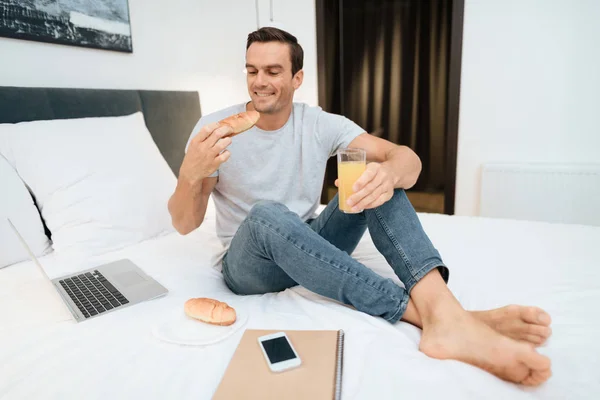 Мужчина сидит на кровати и завтракает. Перед ним стоит открытый серый ноутбук . — стоковое фото