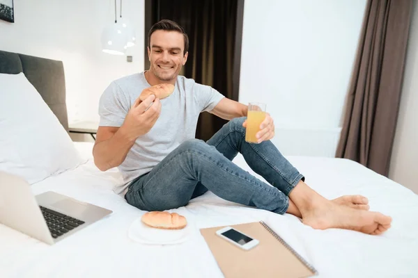 男はベッドの上に座って 朝食を持っています 彼の前に開いているグレー ノート パソコンを略します 彼は寝室で大きな白いベッドの上に座っています — ストック写真