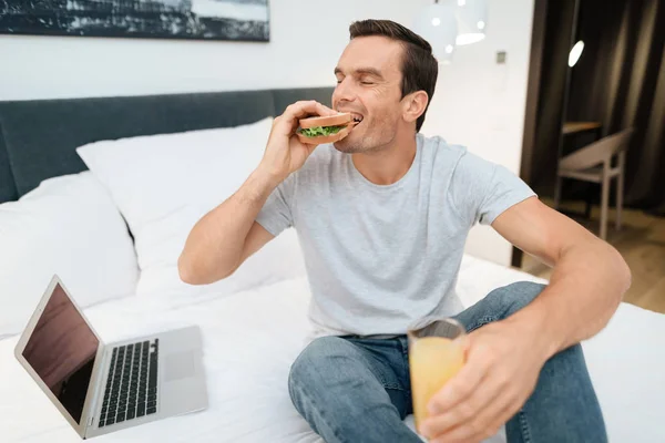 Мужчина сидит на кровати и завтракает. Перед ним стоит открытый серый ноутбук . — стоковое фото