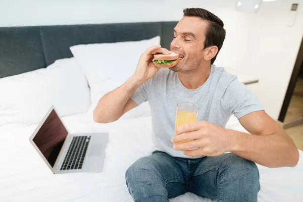 男はベッドの上に座って、朝食を持っています。彼の前に立つオープン グレー ラップトップ. — ストック写真