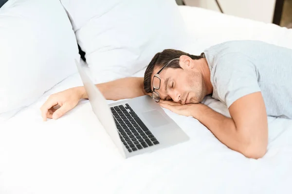 El hombre se quedó dormido en la cama antes de que el portátil estuviera encendido y abierto. Lleva una camiseta gris. . —  Fotos de Stock