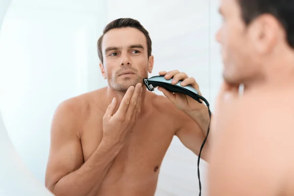 Egy férfi reggel a fürdőszobában. Ő leborotválja a tükör előtt elektromos borotva. — Stock Fotó