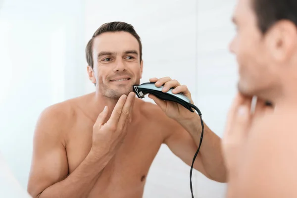 A man is in the bathroom in the morning. He shaves the electric shaver in front of the mirror. A man is in a bathroom with white walls and looks at himself.