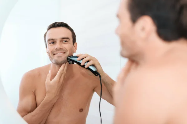 A man in the bathroom in the morning. He shaves the electric shaver in front of the mirror.