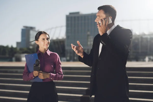 Una chica con una carpeta y un micrófono está esperando que el hombre deje de hablar por teléfono. — Foto de Stock