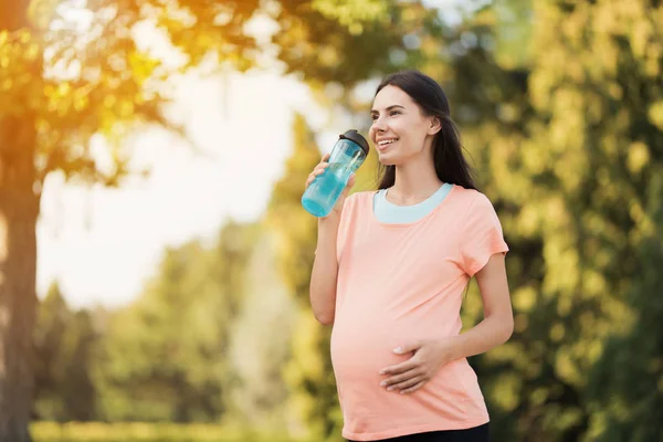 Gravid kvinna promenader i parken med en sport flaska i händerna. Hon dricker ur en flaska — Stockfoto