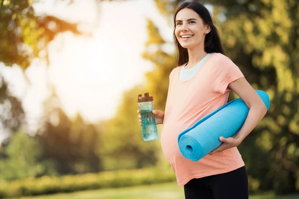 在公园里散步的孕妇手里拿着一个运动瓶子。她有一个瑜伽垫 — 图库照片