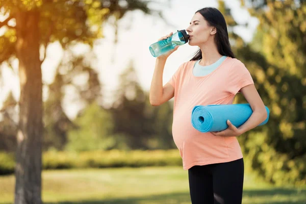 在公园里散步的孕妇手里拿着一个运动瓶子。她从瓶子里喝。她有一个瑜伽垫 — 图库照片