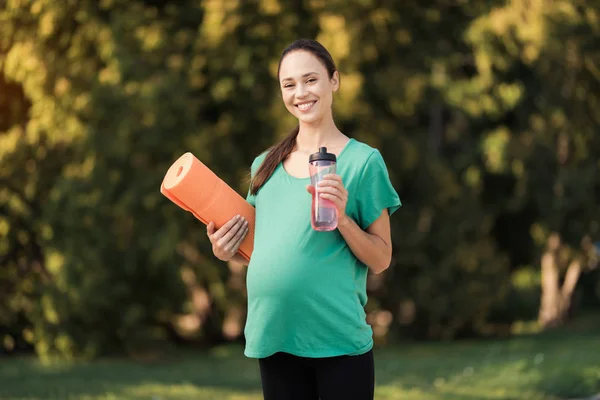 Беременная женщина, гуляющая в парке с бутылкой в руках. У неё коврик для йоги. — стоковое фото