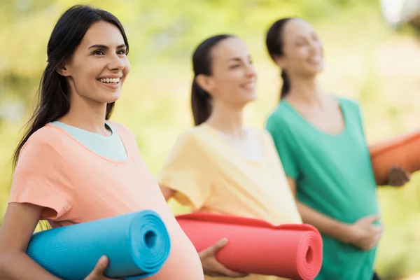 Drei schwangere Mädchen posieren mit Yogamatten in der Hand in einem Park. sie lächeln und haben Spaß — Stockfoto