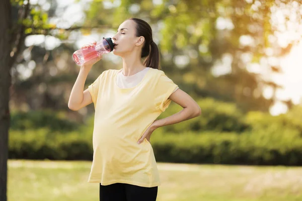 Zwangere vrouw wandelen in het park met een sportfles in handen. Ze drinkt uit een fles — Stockfoto