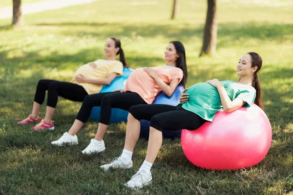 Drei schwangere Frauen liegen auf Yoga-Bällen im Park. sie halten ihre Bäuche — Stockfoto