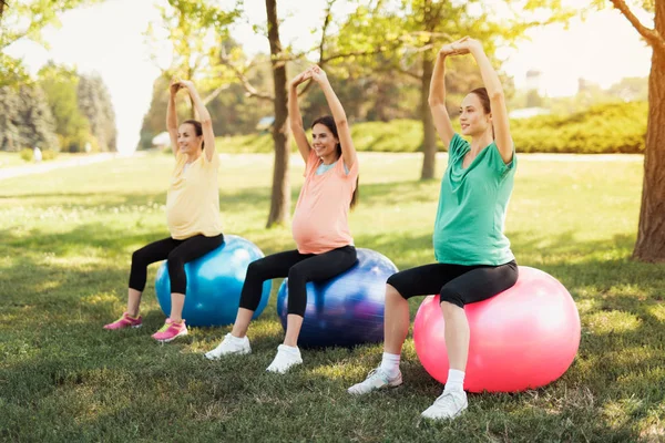 Drei schwangere Frauen sitzen mit erhobenen Händen in einem Park auf Yoga-Bällen — Stockfoto