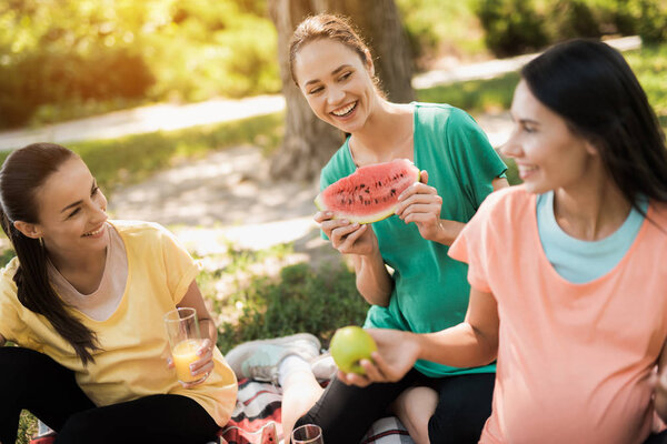 Three pregnant women sit in the park on a rug for picnics and eat. They are all smiling