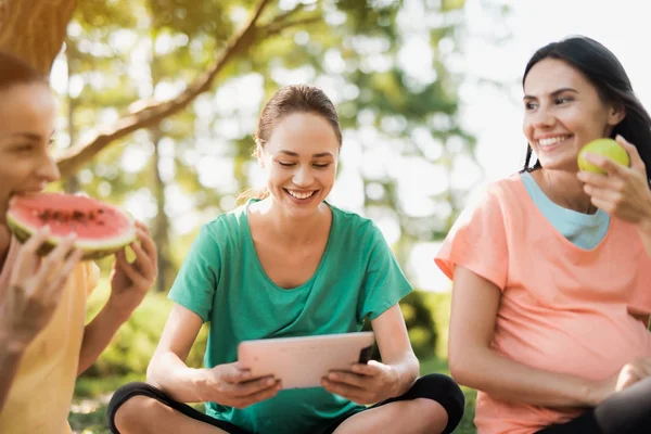 Tres mujeres embarazadas descansan en la naturaleza después de hacer yoga. Uno de ellos sostiene una tableta — Foto de Stock