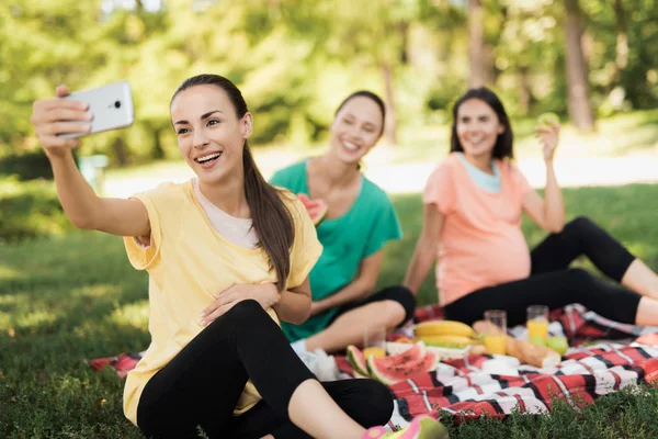 Kobiety w ciąży w żółty T-shirt siedzi z koleżankami w ciąży na piknik w parku i sprawia, że selfie — Zdjęcie stockowe