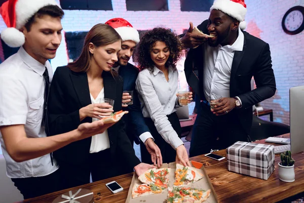 People are preparing to meet the new year in the office. They hold glasses with champagne and pizza.