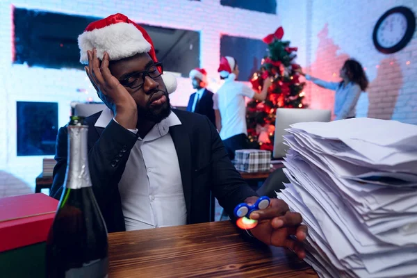 Un uomo di colore è seduto alla sua scrivania la vigilia di Capodanno. Ha un sacco di lavoro e gira lo spinner dalla noia . — Foto Stock