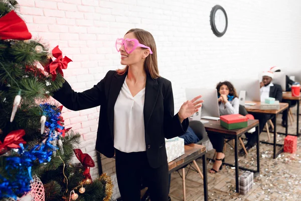 Ein Mädchen im strengen Anzug kleidet einen Weihnachtsbaum. Sie trägt eine rosafarbene Brille und lächelt. — Stockfoto