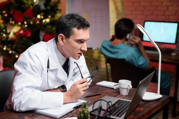 Il dottore lavora la vigilia di Capodanno. Sta parlando con qualcuno in video. Dietro di lui lavora un collega . — Foto Stock