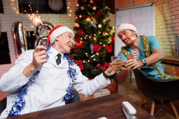 Due medici celebrano il nuovo anno in ufficio. Essi battono i bicchieri con whisky e luci bergina nelle loro mani . — Foto Stock