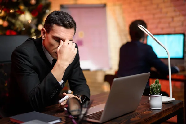 Empresario sentado en la oficina y trabajando en la víspera de Año Nuevo. Cerró los ojos y se aferró al puente de su nariz. . — Foto de Stock