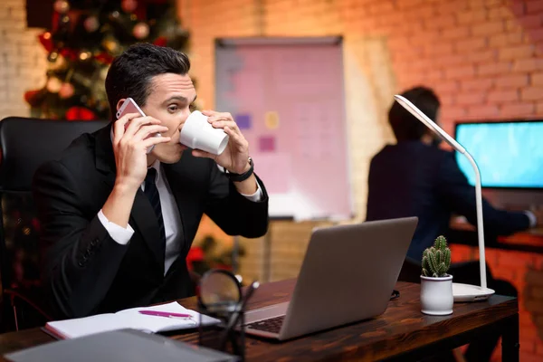 Homme d'affaires assis au bureau et travaillant la veille du Nouvel An. Il parle au téléphone et boit du café dans la tasse . — Photo