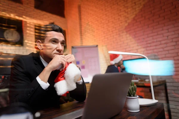 Uomo d'affari seduto in ufficio e che lavora alla vigilia di Capodanno. Guarda pensieroso da parte e tiene il berretto di un Anno nuovo — Foto Stock