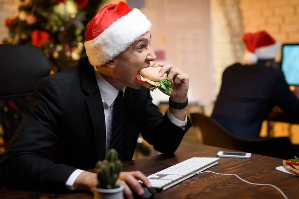 Uomo d'affari in ufficio la vigilia di Capodanno. Mangia un hamburger e guarda qualcosa sul computer. . — Foto Stock