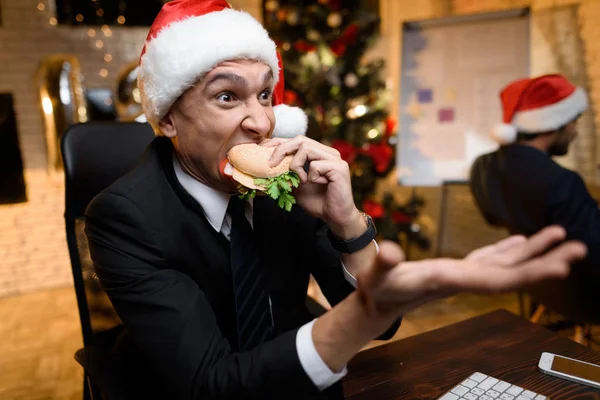 Uomo d'affari in ufficio la vigilia di Capodanno. Mangia un hamburger e guarda qualcosa sul computer. . — Foto Stock