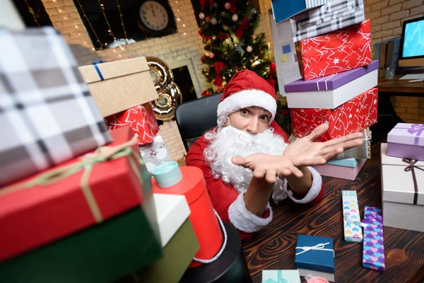 Podnikatel v kanceláři na Silvestra. On nosí Santa Claus kostým. Kolem něj hodně dárků, — Stock fotografie