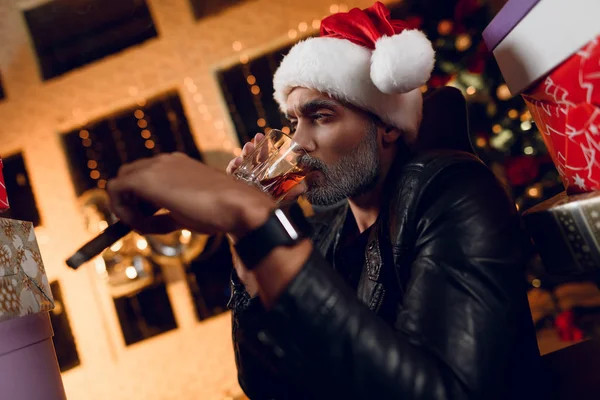 Elegante Babbo Natale in una giacca di pelle nera fuma un sigaro e beve whisky. Si siede circondato dai regali di Capodanno . — Foto Stock