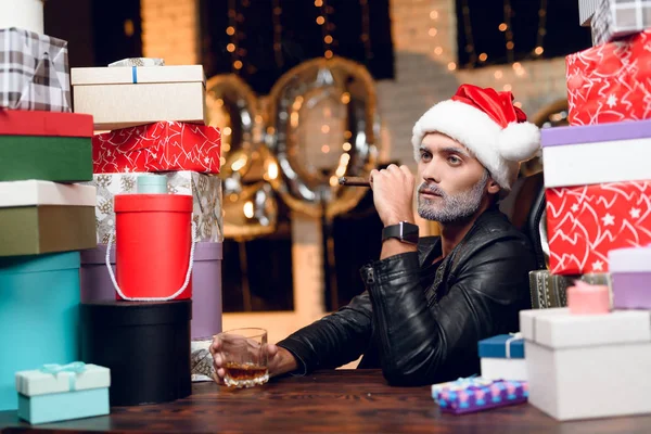 Elegante Babbo Natale in una giacca di pelle nera fuma un sigaro e beve whisky. Si siede circondato dai regali di Capodanno . — Foto Stock