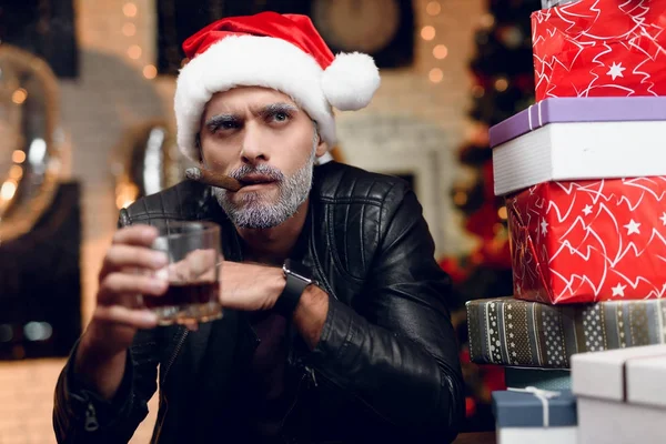 Brutal Papai Noel em uma jaqueta de couro preto senta-se com um charuto em sua boca e um copo de uísque em suas mãos — Fotografia de Stock