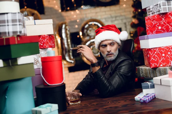 Elegante Babbo Natale in una giacca di pelle nera fuma un sigaro e beve whisky. Si siede circondato dai regali di Capodanno . — Foto Stock