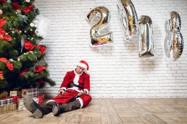 Um homem bêbado no traje do Papai Noel adormeceu perto da árvore de Ano Novo com uma garrafa de champanhe e um charuto nas mãos. . — Fotografia de Stock