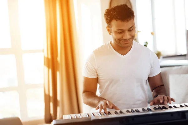 Un hombre negro se sienta en la sala de su apartamento y toca un sintetizador. Compone música. . — Foto de Stock