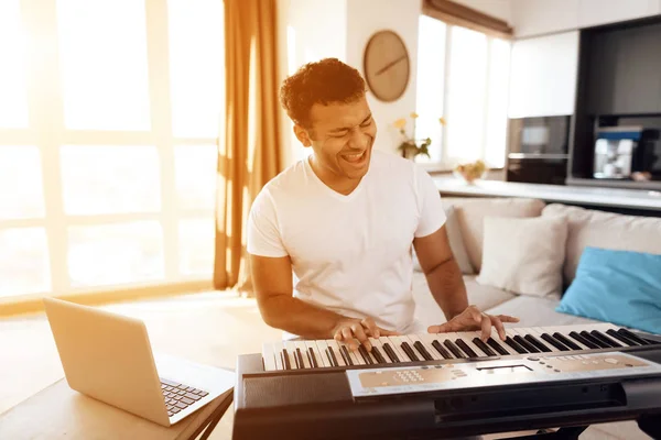 Чорний чоловік сидить у вітальні своєї квартири і грає синтезатора. Він створює музику . — стокове фото