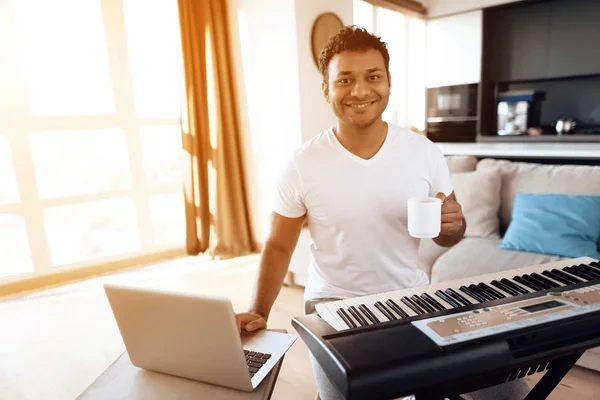 Un hombre negro se sienta en la sala de su apartamento y toca un sintetizador. Compone música y bebe café . —  Fotos de Stock