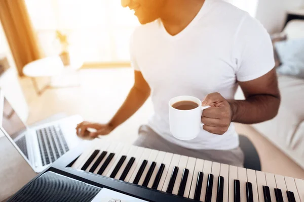 Un uomo di colore siede nel soggiorno del suo appartamento e suona un sintetizzatore. Compone musica e beve caffè . — Foto Stock