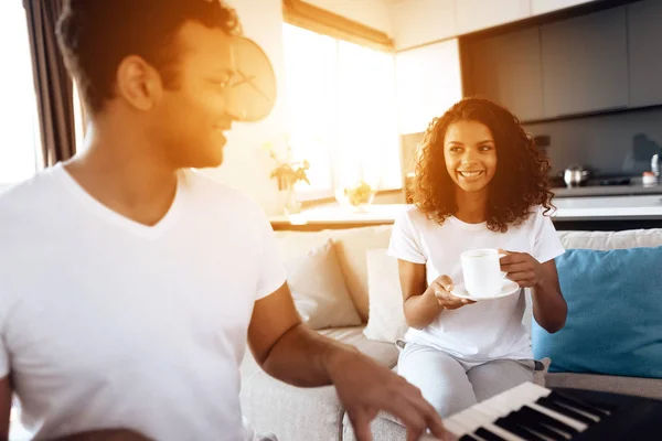 Черный мужчина сидит в гостиной своей квартиры и играет на синтезаторе. Девушка, сидящая рядом с ним и пьющая чай . — стоковое фото
