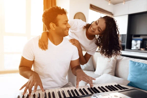 Черный мужчина сидит в гостиной своей квартиры и играет на синтезаторе. Рядом его девушка и обнимает его. . — стоковое фото